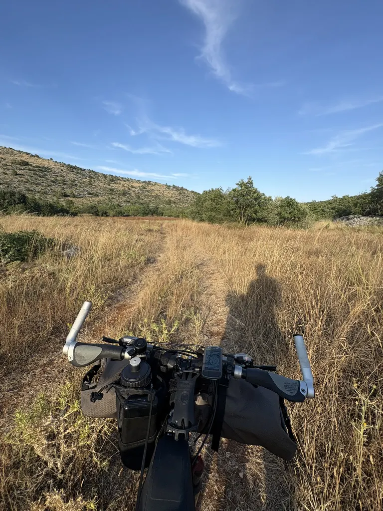 Handlebar view of a faint track running through waist-high dry grass towards low scrub hills.