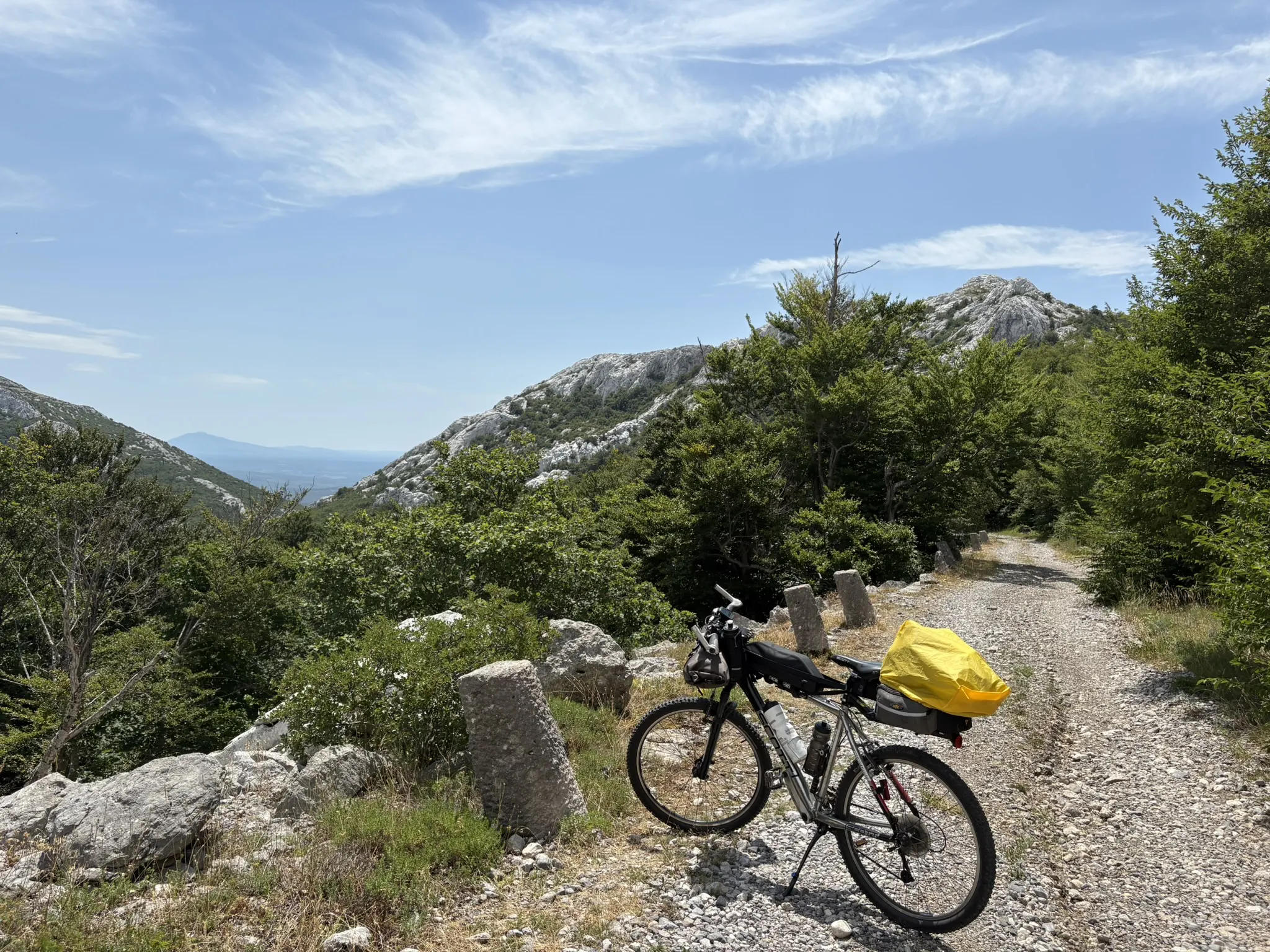 The bike with a yellow drybag parked on a gravel track through karst limestone and green trees.