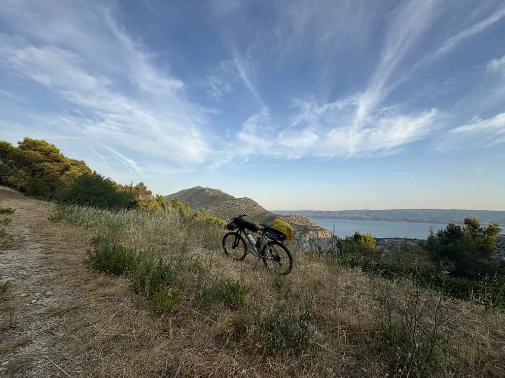 A loaded touring bike propped on a grassy ridge above a bay, hills and thin cirrus cloud beyond.