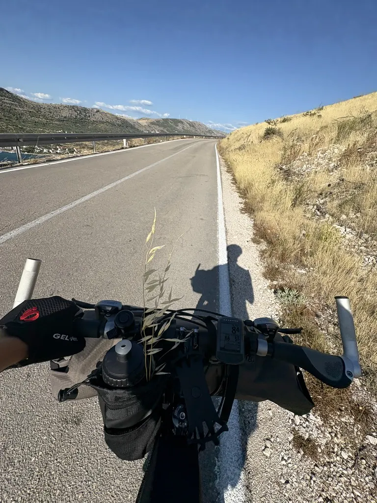 Handlebar view along the white line of a coastal road, bar bag and cycling computer in frame, dry hillside to the right.