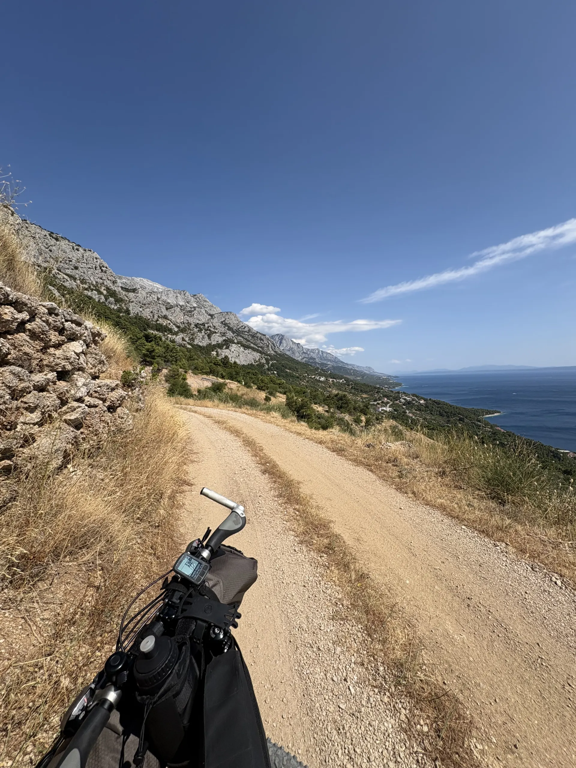 A gravel road curving down a steep hillside towards the sea, mountains dropping to the coast.