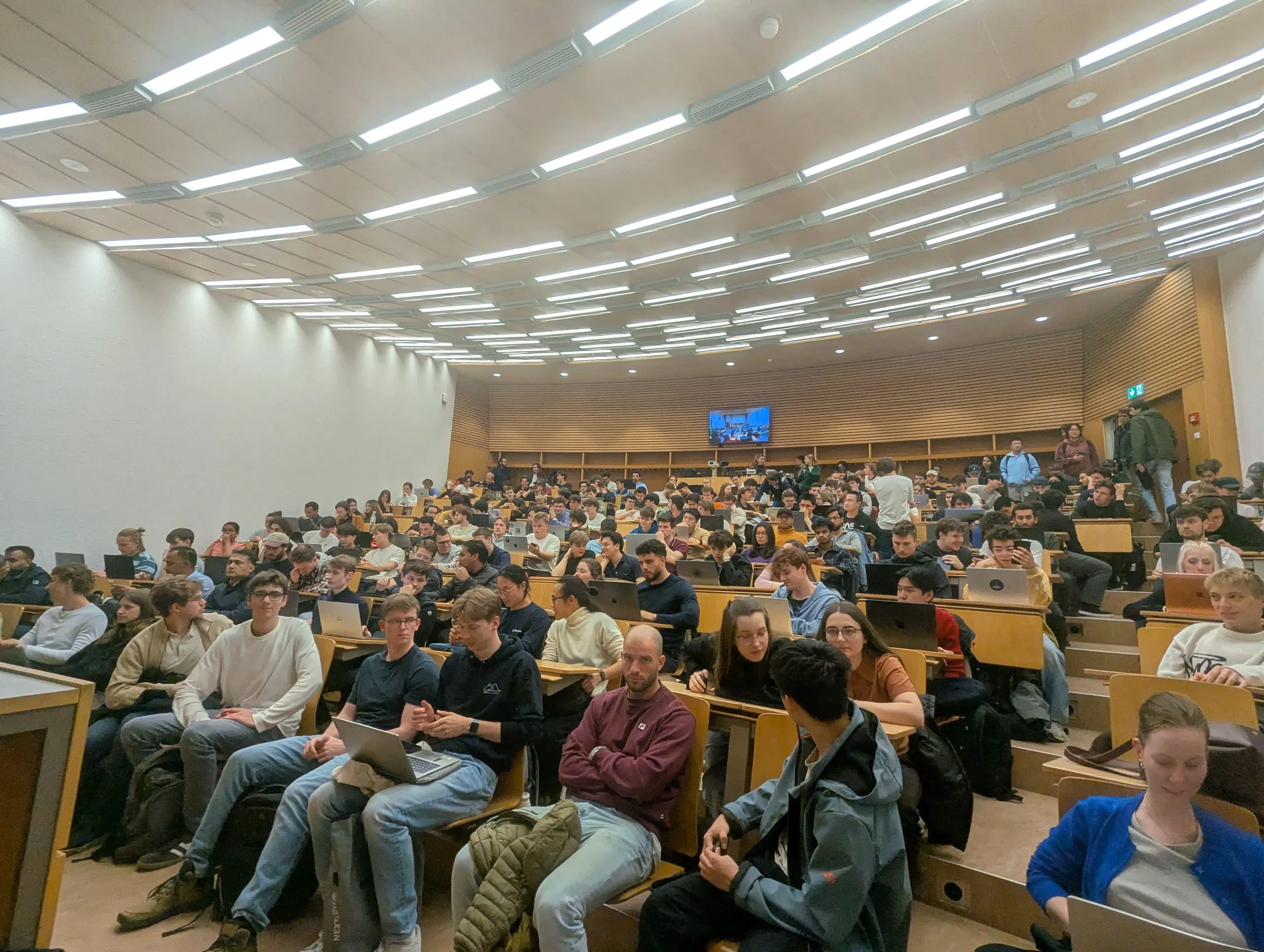 The lecture hall seen from the upper back rows, every desk taken, laptops open along the tiered benches.